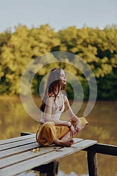 A young happy woman with a hippie smile sits on the lake shore on a bridge wearing eco clothing made of natural