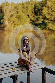 A young happy woman with a hippie smile sits on the lake shore on a bridge wearing eco clothing made of natural