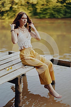 A young happy woman with a hippie smile sits on the lake shore on a bridge wearing eco clothing made of natural