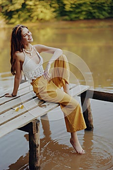 A young happy woman with a hippie smile sits on the lake shore on a bridge wearing eco clothing made of natural