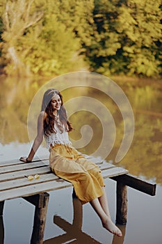 A young happy woman with a hippie smile sits on the lake shore on a bridge wearing eco clothing made of natural