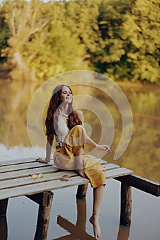 A young happy woman with a hippie smile sits on the lake shore on a bridge wearing eco clothing made of natural