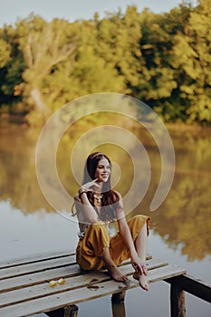 A young happy woman with a hippie smile sits on the lake shore on a bridge wearing eco clothing made of natural