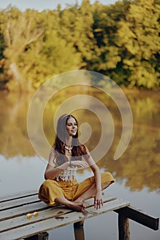 A young happy woman with a hippie smile sits on the lake shore on a bridge wearing eco clothing made of natural