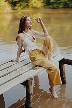 A young happy woman with a hippie smile sits on the lake shore on a bridge wearing eco clothing made of natural