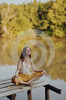 A young happy woman with a hippie smile sits on the lake shore on a bridge wearing eco clothing made of natural