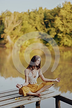 A young happy woman with a hippie smile sits on the lake shore on a bridge wearing eco clothing made of natural