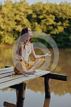 A young happy woman with a hippie smile sits on the lake shore on a bridge wearing eco clothing made of natural