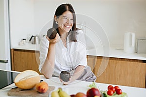 Young happy woman holding avocado in hand and smiling on background of fresh fruits and vegetables in modern white kitchen.