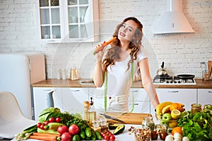 Young happy woman holding carrot in the beautiful kitchen with green fresh ingredients indoors. Healthy food and Dieting concept.