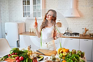 Young happy woman holding carrot in the beautiful kitchen with green fresh ingredients indoors. Healthy food and Dieting concept.