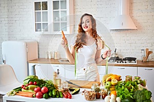 Young happy woman holding carrot in the beautiful kitchen with green fresh ingredients indoors. Healthy food and Dieting concept.