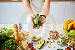 Young happy woman holding cucumbers for making salad in the beautiful kitchen with green fresh ingredients indoors. Healthy food