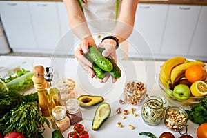 Young happy woman holding cucumbers for making salad in the beautiful kitchen with green fresh ingredients indoors. Healthy food