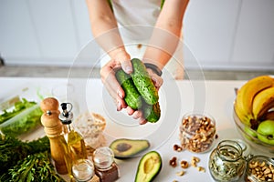 Young happy woman holding cucumbers for making salad in the beautiful kitchen with green fresh ingredients indoors. Healthy food