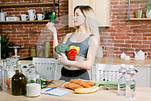 Young and happy woman holding frech cucumber and bowl with fresh healthy vegetables standing on the table with rice