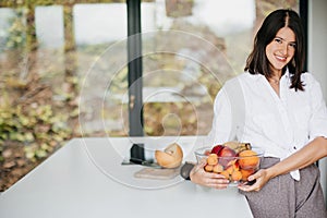 Young happy woman holding glass bowl with summer fruits on background of modern white kitchen. Healthy food concept. Home cooking