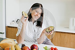 Young happy woman holding halves of ripe avocado and smiling in modern kitchen. Healthy eating