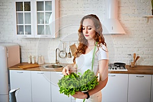 Young happy woman holding lettuce leaves for making salad in the beautiful kitchen with green fresh ingredients indoors. Healthy