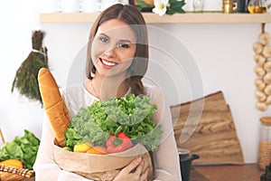 Young happy woman holding paper bag full of vegetables and fruits while smiling. Girl have made shopping and ready for
