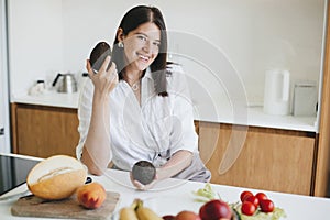 Young happy woman holding ripe avocado in hands and smiling in modern white kitchen. Healthy eating