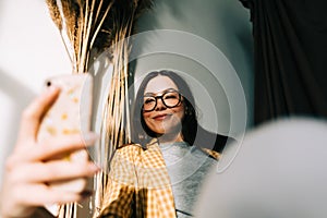 Young happy woman holding smartphone looking on cell using mobile phone technology at home, taking selfie for social media or