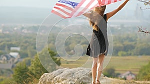 Young happy woman with long hair raising up waving on wind american national flag in her hands climbing on high rocky hill enjoyin