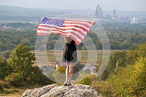 Young happy woman with long hair raising up waving on wind american national flag in her hands standing on high rocky hill