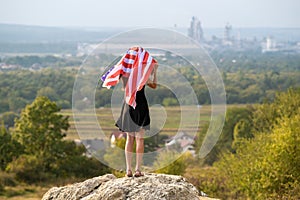 Young happy woman with long hair raising up waving on wind american national flag in her hands standing on high rocky hill