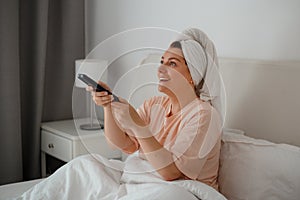 Young happy woman lying on bed and watching television. Positive smiling girl sit indoors at home watch tv holding