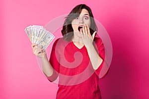 Young happy woman with money in hand, with opened mouth, looks surprised. Brunette girl wins in lottery. Lucky female dressed red