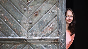 Young happy woman peeks out from behind big ancient doors.