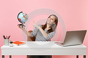 Young happy woman pointing on globe, planning vacation while sit, work at office at white desk with contemporary pc