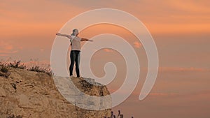 Young happy woman raises her hands up, standing on top of a mountain above sea against backdrop of blue sky. Watching