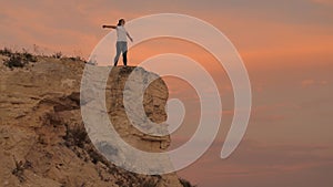 Young happy woman raises her hands up, standing on top of a mountain above sea against backdrop of blue sky. Watching