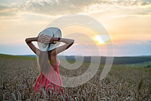Young happy woman in red summer dress and white straw hat standing on yellow farm field with ripe golden wheat enjoying warm
