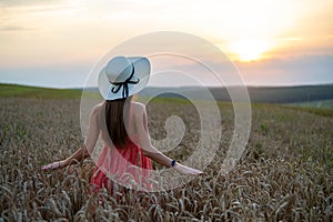 Young happy woman in red summer dress and white straw hat standing on yellow farm field with ripe golden wheat enjoying warm