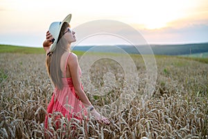 Young happy woman in red summer dress and white straw hat standing on yellow farm field with ripe golden wheat enjoying warm