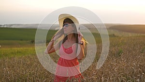 Young happy woman in red summer dress and white straw hat walking on yellow farm field with ripe golden wheat enjoying