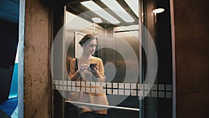 Young happy woman riding elevator with glass wall, door opens and she walks out using smartphone mobile shopping app.