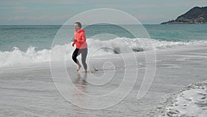 Young happy woman is running barefoot on waves. Attractive female is jogging on sandy beach, enjoying beautiful seascape. Carefree