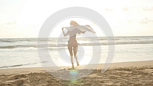Young happy woman with scarf running on the ocean beach at sunset. Girl enjoying summer. Female in bikini with flying