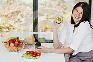 Young happy woman showing half of perfectly ripe avocado in hand and smiling on background of fresh fruits and vegetables in