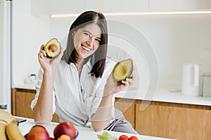 Young happy woman showing half of perfectly ripe avocado in hands and smiling on background of fresh fruits and vegetables in