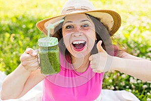 Young happy woman showing thumbs up with green smoothies at a picnic. Healthy food, detox and diet concept