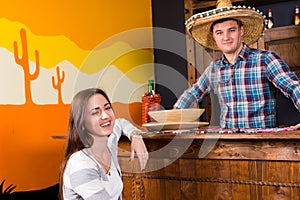 Young happy woman sitting at the bar counter next to the bartender wearing sombrero