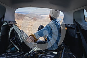 Young happy woman sitting in an open car trunk with her arms outstretched. Travel by car concept