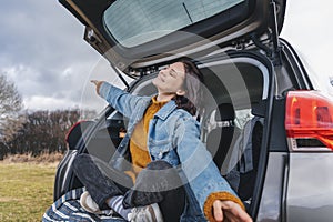 Young happy woman sitting in an open car trunk with her arms outstretched. Travel by car concept