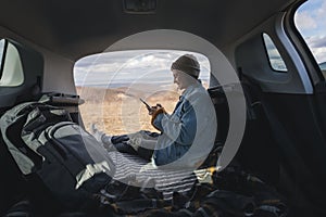 Young happy woman sitting in an open car trunk with a smartphone in her hands. Traveling by car, communication in travel