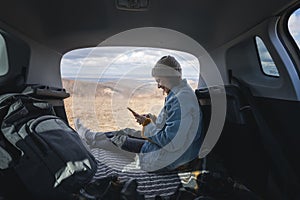 Young happy woman sitting in an open car trunk with a smartphone in her hands. Traveling by car, communication in travel